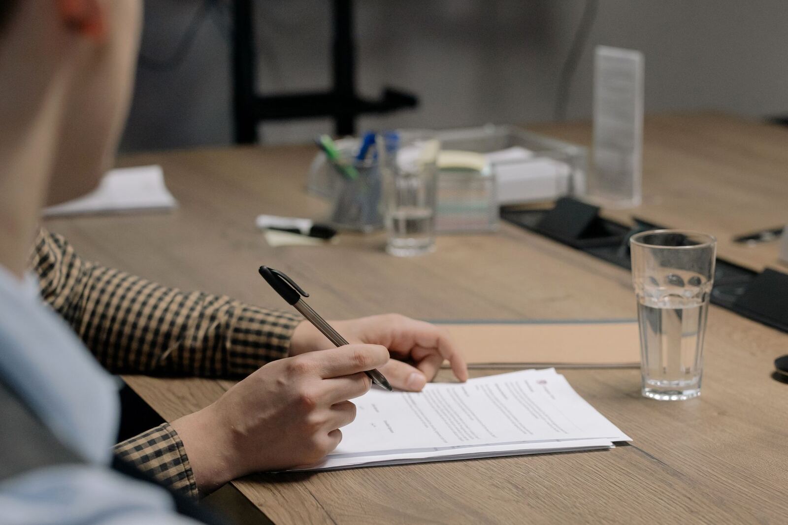 Person in business attire signing a document at a wooden table in an office setting.